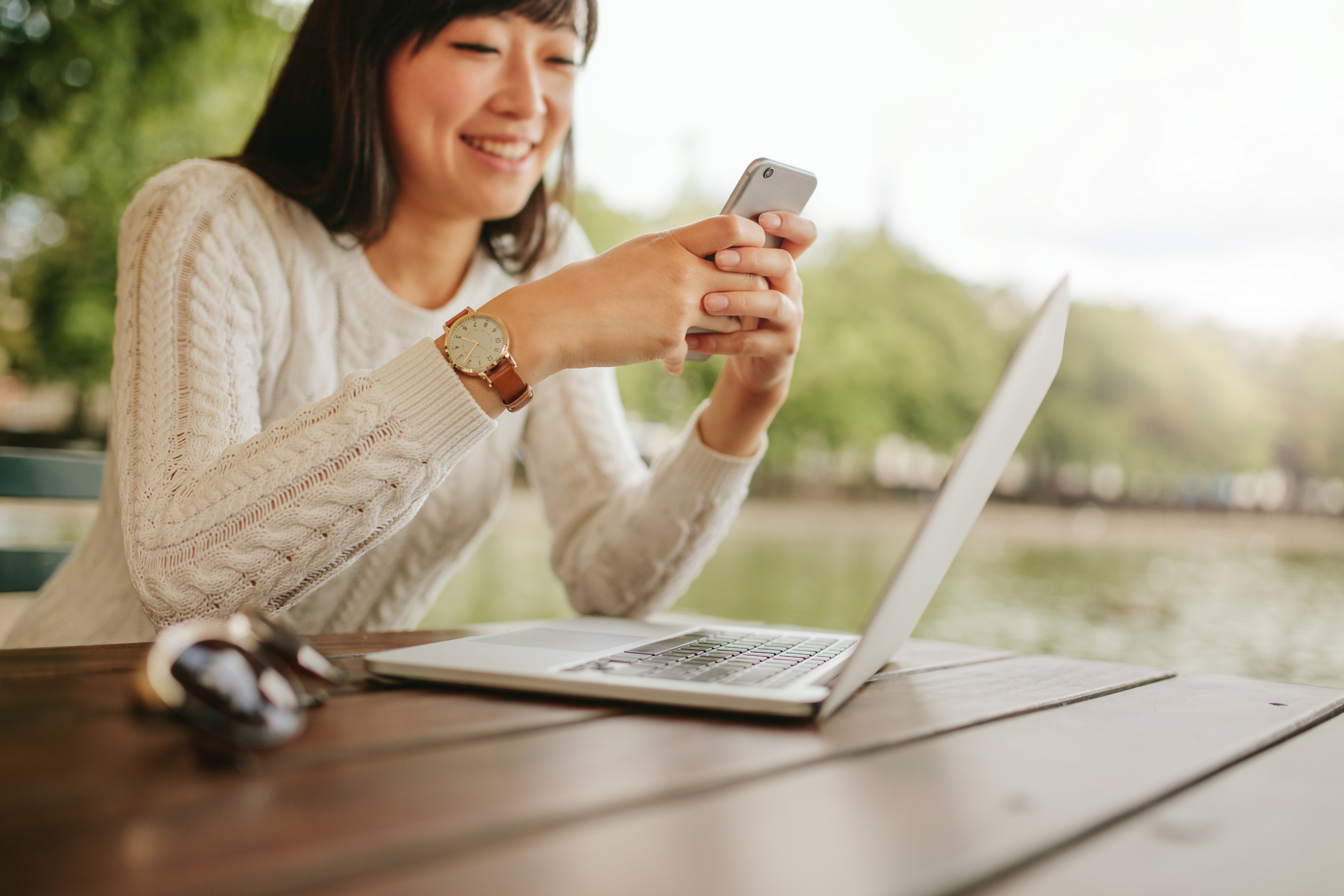 Shot of smiling woman using mobile phone at outdoor cafe. Young female sitting at table with laptop reading text message on her smart phone.