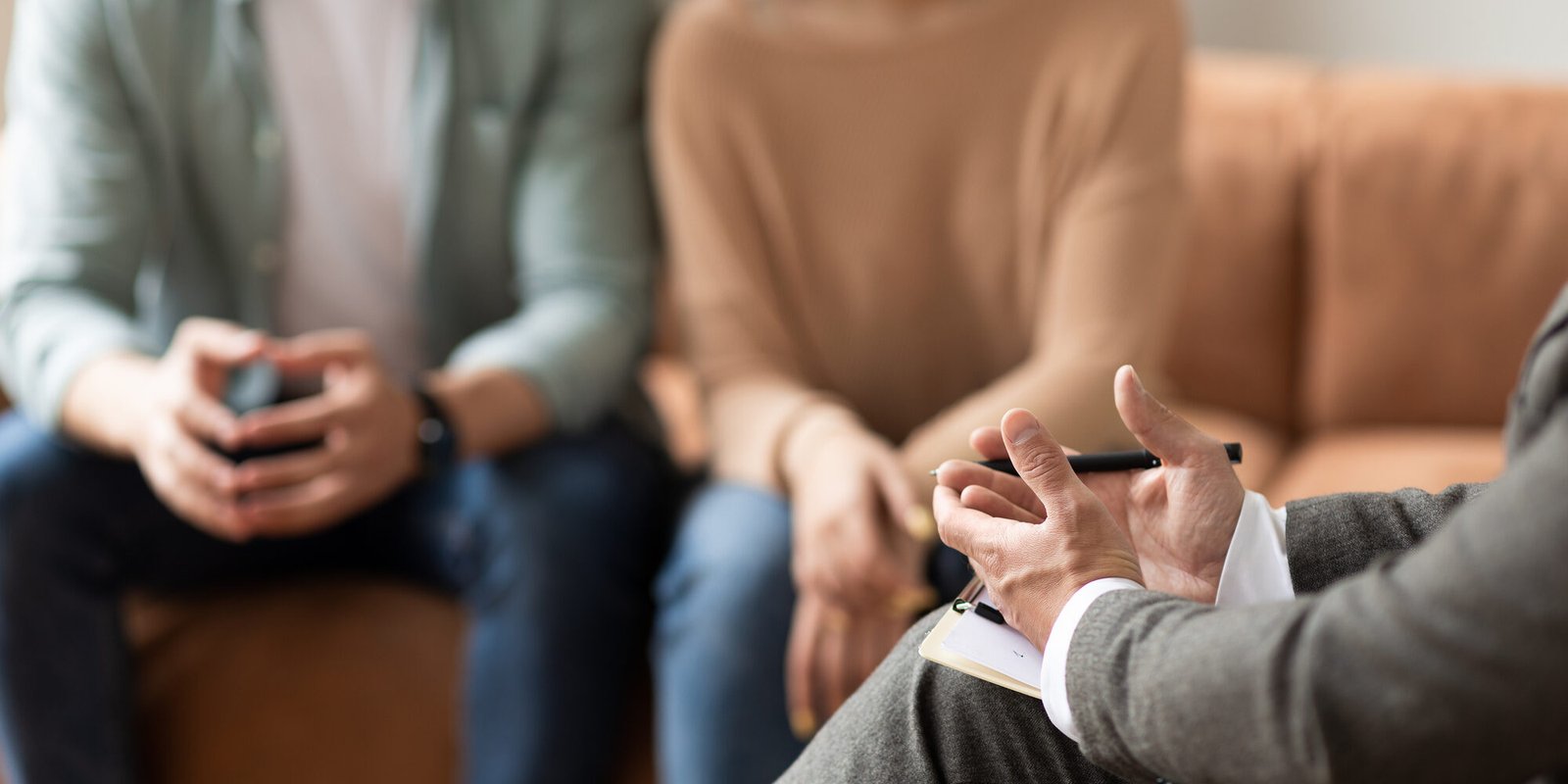 Help Concept. Closeup Cropped View Of Male Counselor Therapist Or Consultant In Suit Talking With Couple Sitting In Office, Giving Professional Advice. Selective Focus On Hands With Clipboard And Pen