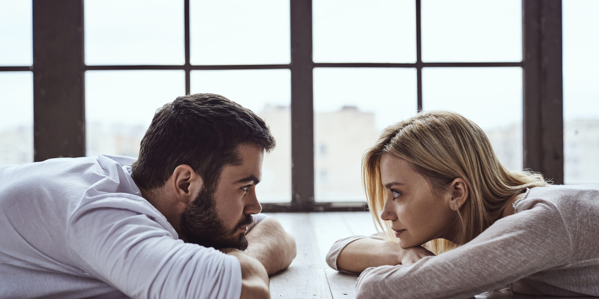 Young happy loving couple guy and girl lying against each other in front of big panoramic window and looking at each other