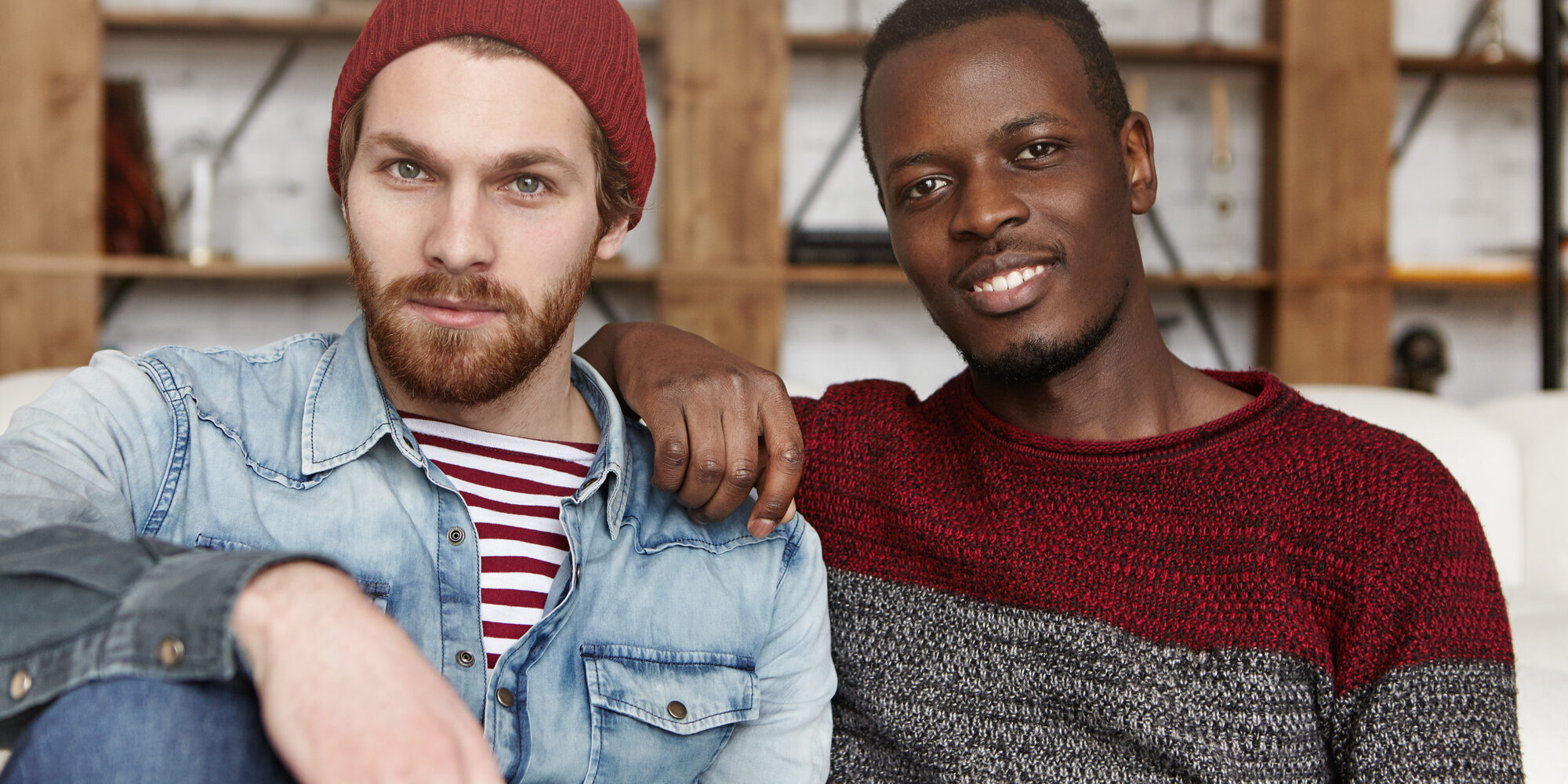 Homosexual love and relationships concept. Interracial gay couple relaxing at cafe: African-American man in sweater holding hand on his stylish bearded Caucasian boyfriend's shoulder in trendy hat
