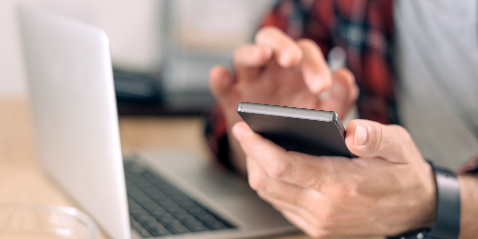 Usando um smartphone e um laptop no escritório em casa, feche as mãos masculinas, usando eletrônicos modernos para teletrabalho, foco seletivo